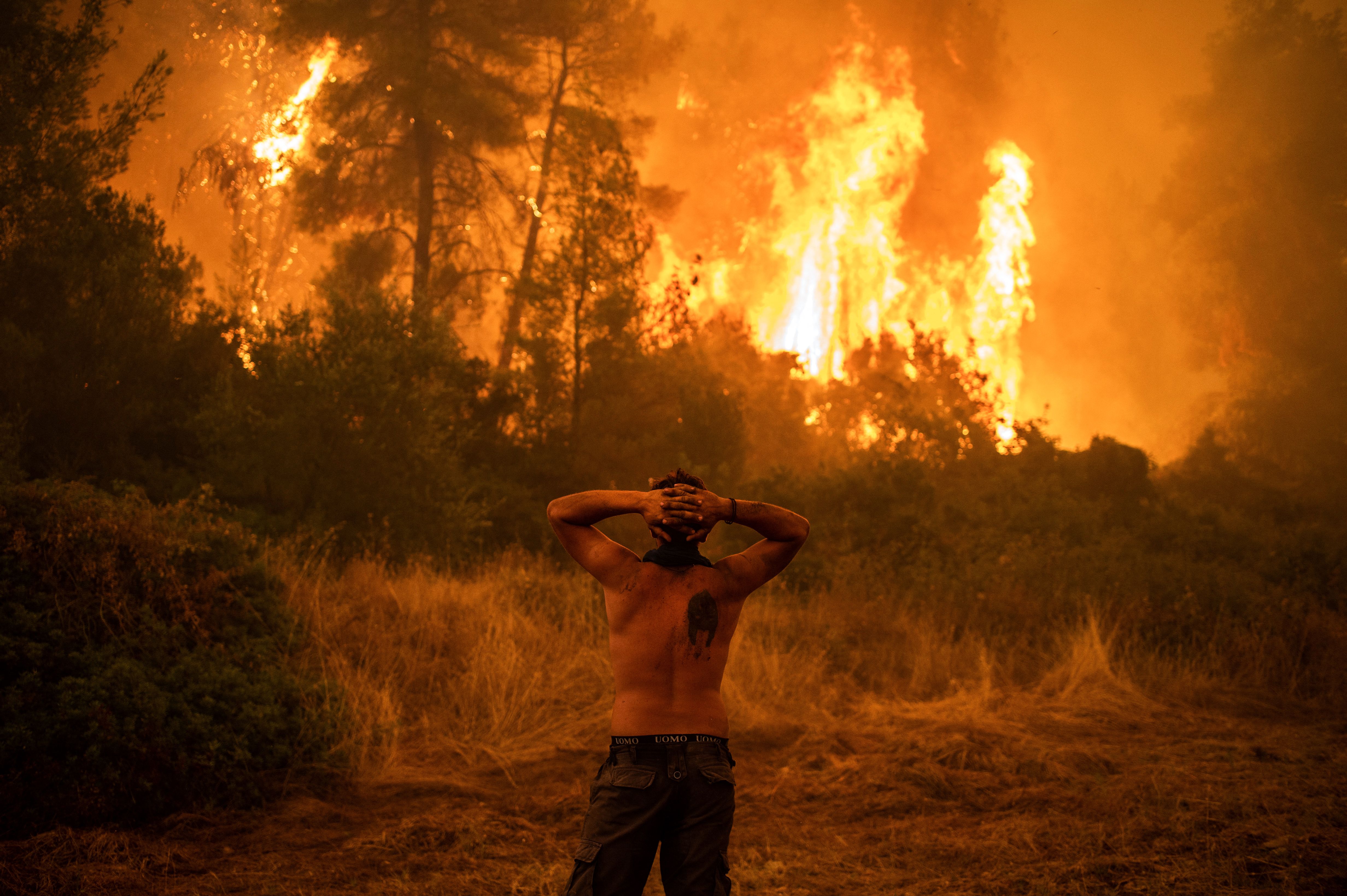Ein oberkörperfreier Mann blickt mit hinter dem Kopf verschränkten Händen auf einen Waldbrand